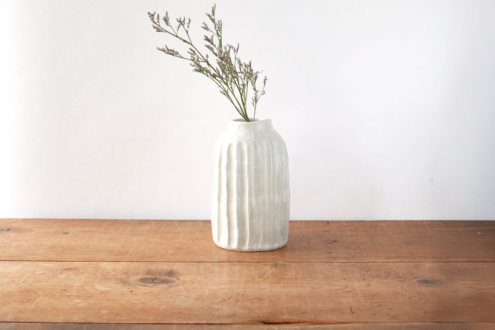 A white ribbed ceramic vase with a slightly flared mouth, filled with small flowers, standing on a rustic wooden tabletop against a plain white wall.