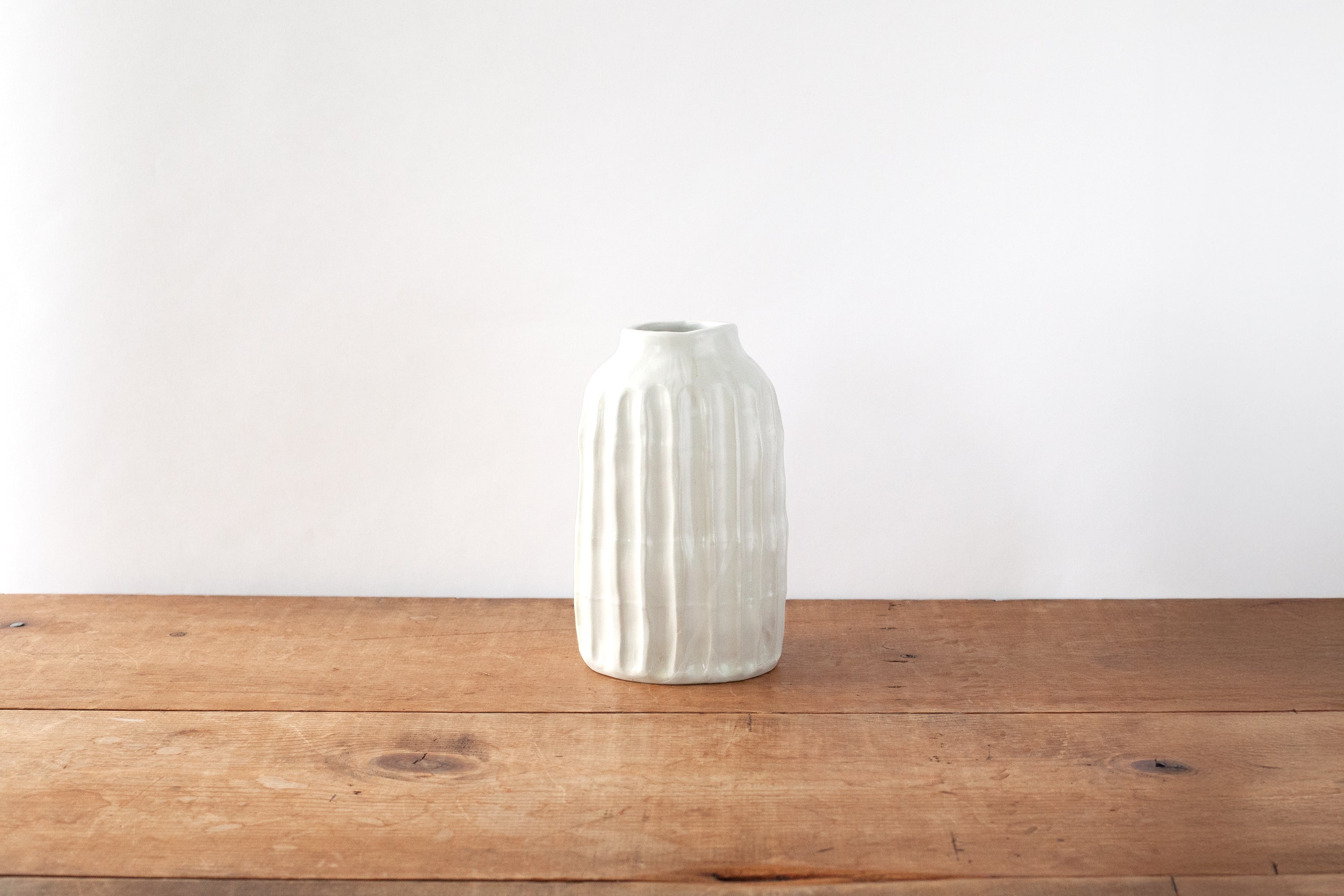 A white ribbed ceramic vase with a slightly flared mouth, standing on a rustic wooden tabletop against a plain white wall.