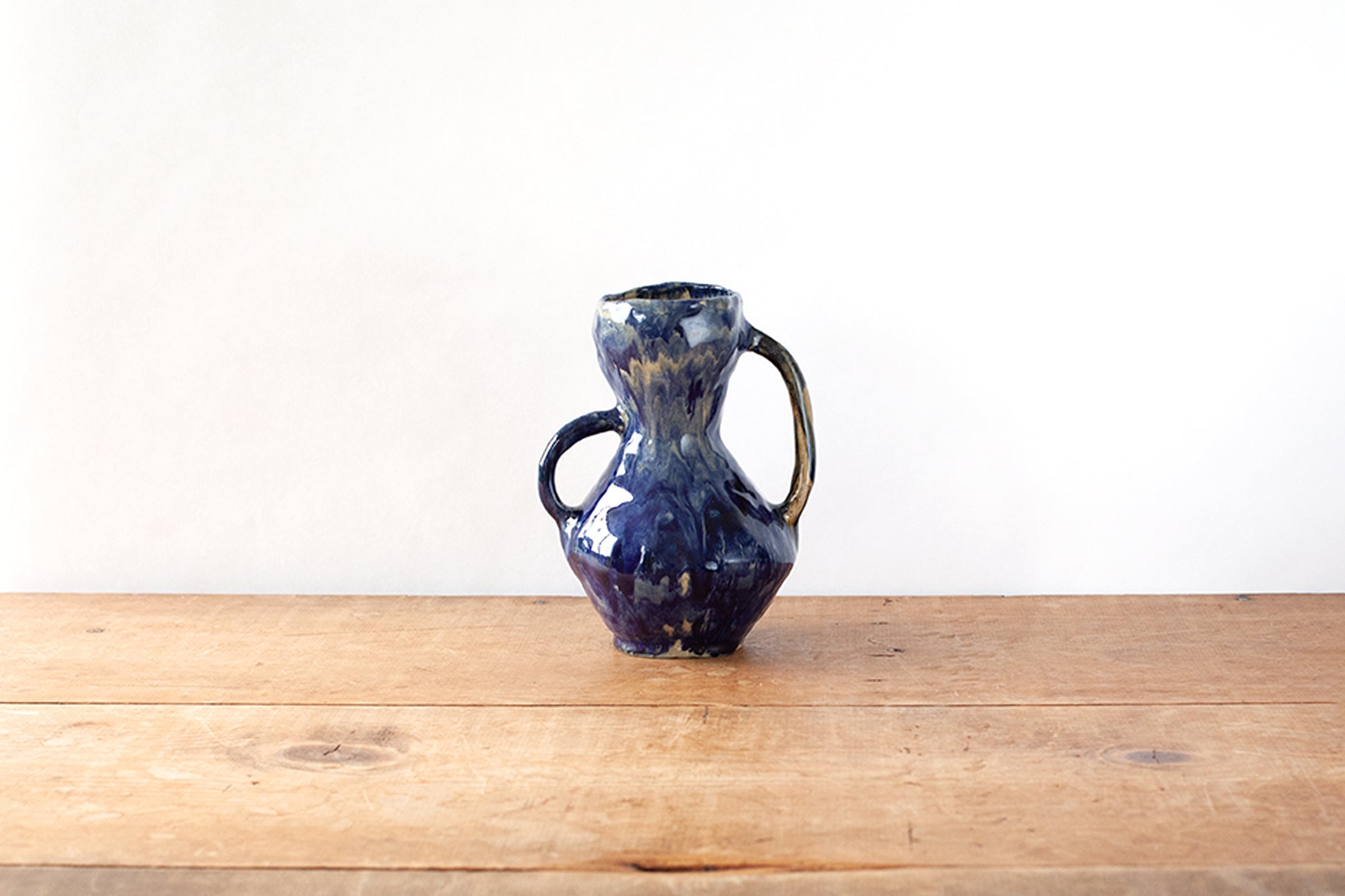 A small handmade ceramic handled vase with a dark glossy blue glaze, sitting on a wooden tabletop against a plain white wall.