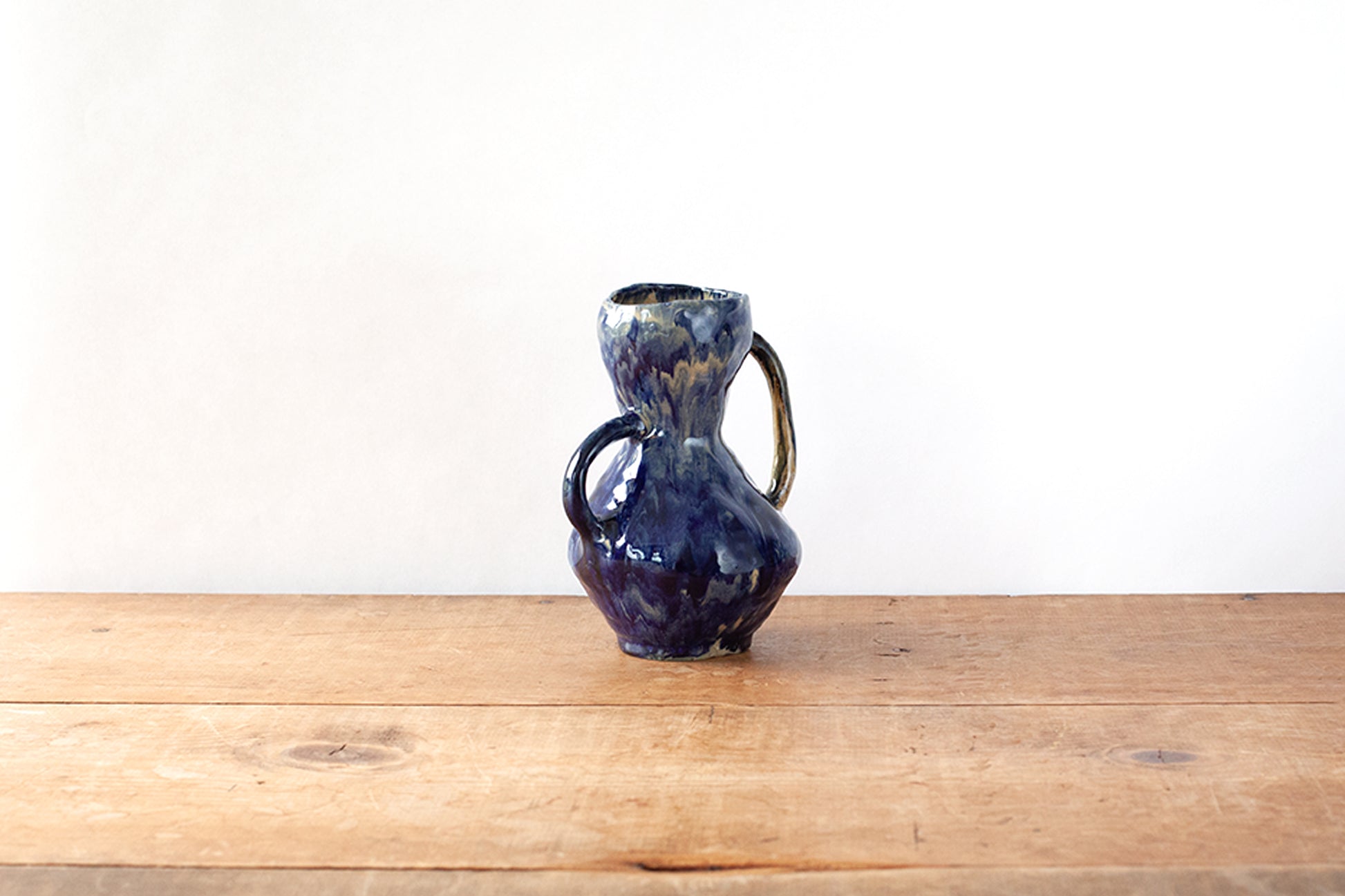 A small handmade ceramic handled vase with a dark glossy blue glaze, sitting on a wooden tabletop against a plain white wall.