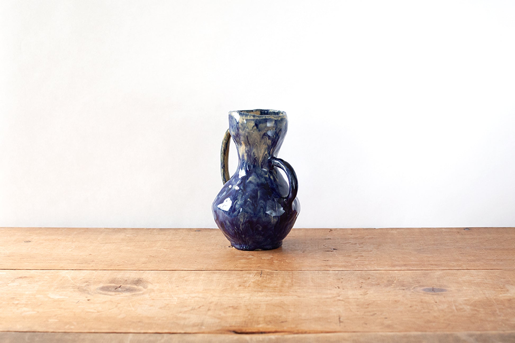 A small handmade ceramic handled vase with a dark glossy blue glaze, sitting on a wooden tabletop against a plain white wall.