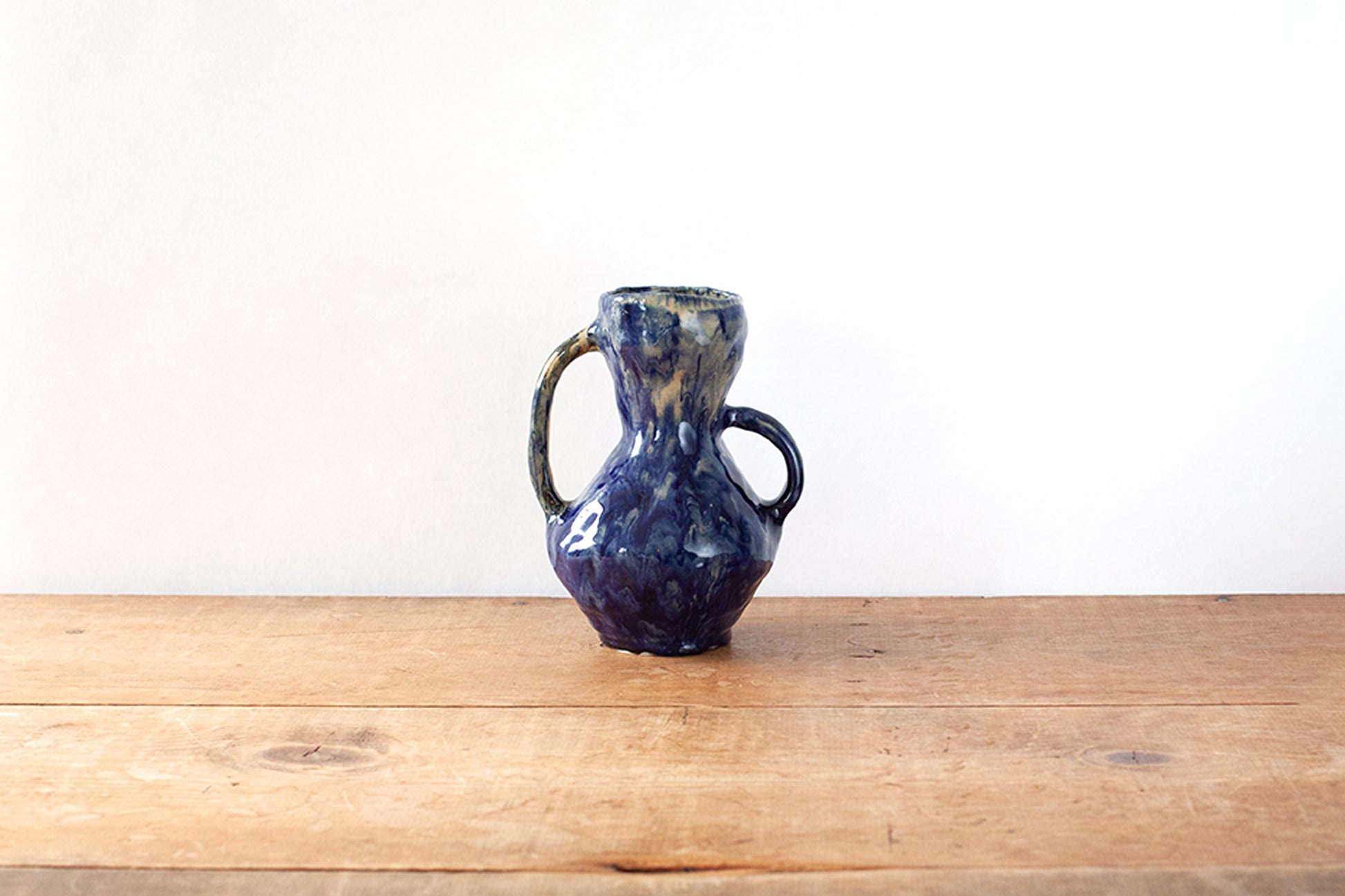A small handmade ceramic handled vase with a dark glossy blue glaze, sitting on a wooden tabletop against a plain white wall.