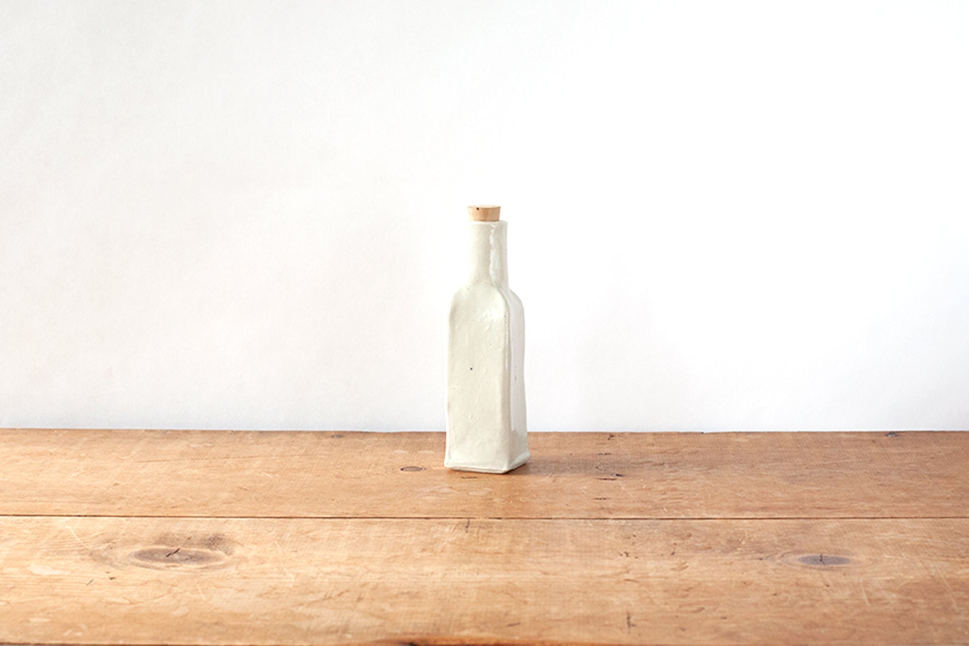 A small off-white ceramic bottle with a matching cork stopper, standing on a rustic wooden tabletop against a plain white wall.