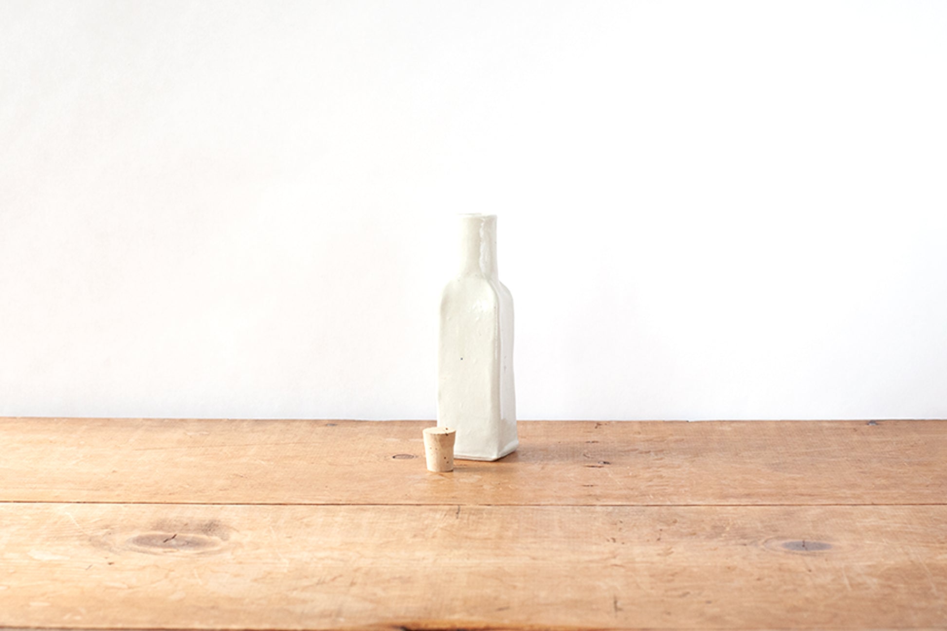 A small off-white ceramic bottle with a matching cork stopper, standing on a rustic wooden tabletop against a plain white wall.