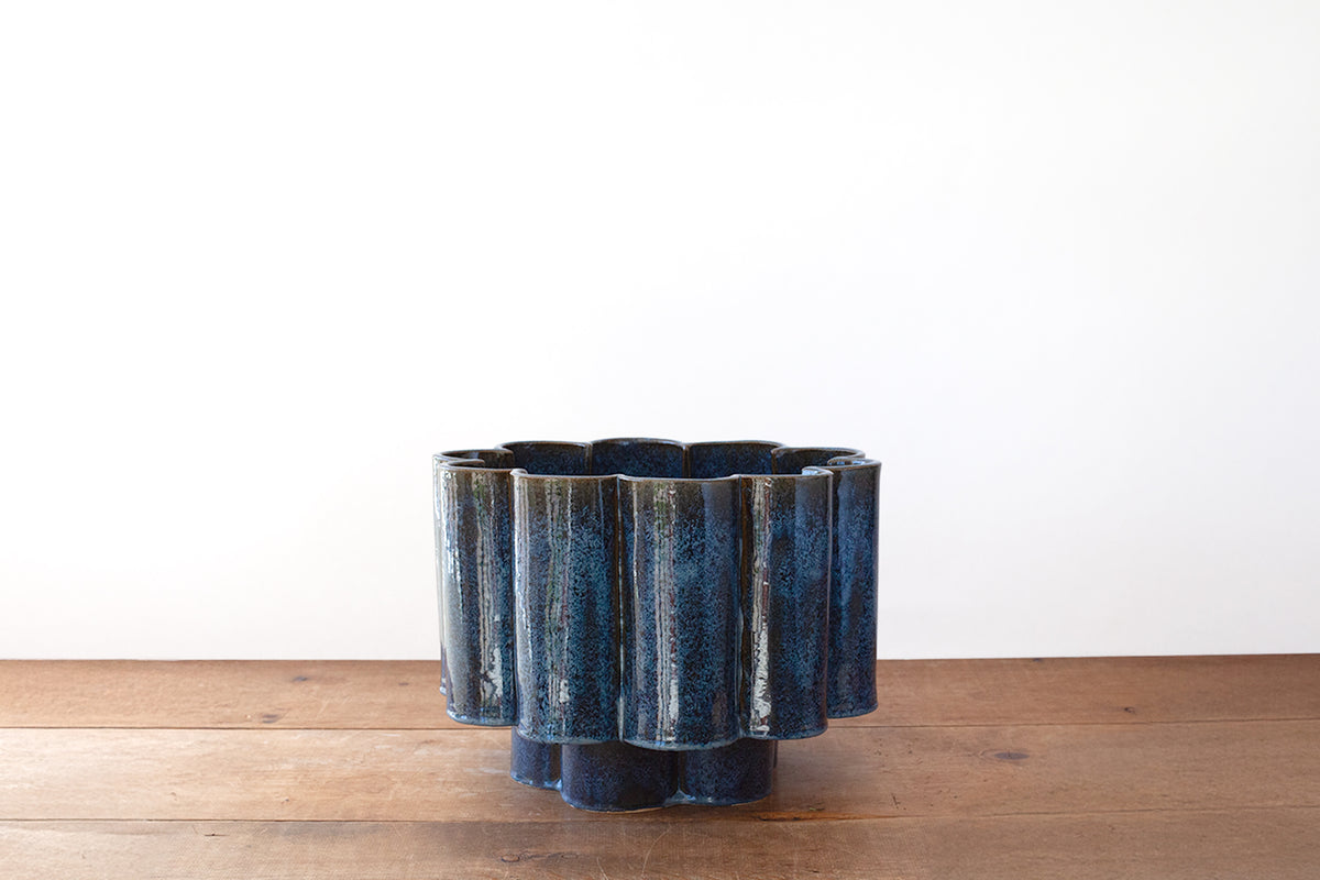 A sculptural ceramic planter with a scalloped design, featuring a mottled dark blue glaze, set on a wooden surface against a plain white wall.