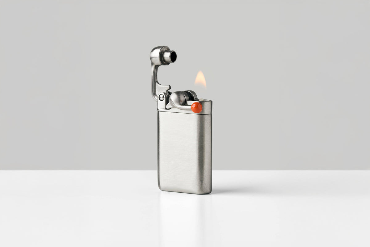 A brushed stainless steel pocket lighter with a lit flame, standing on a white surface against a light gray background.