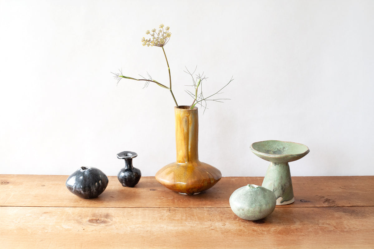 A collection of handmade ceramic vases arranged on a rustic wooden table against a plain white wall. The central, most prominent piece is a tall, narrow mustard-yellow glazed vase holding a dried flower stem surrounded by a small black vase, a round sage-green bowl stand, and a round sage-green ceramic orb.