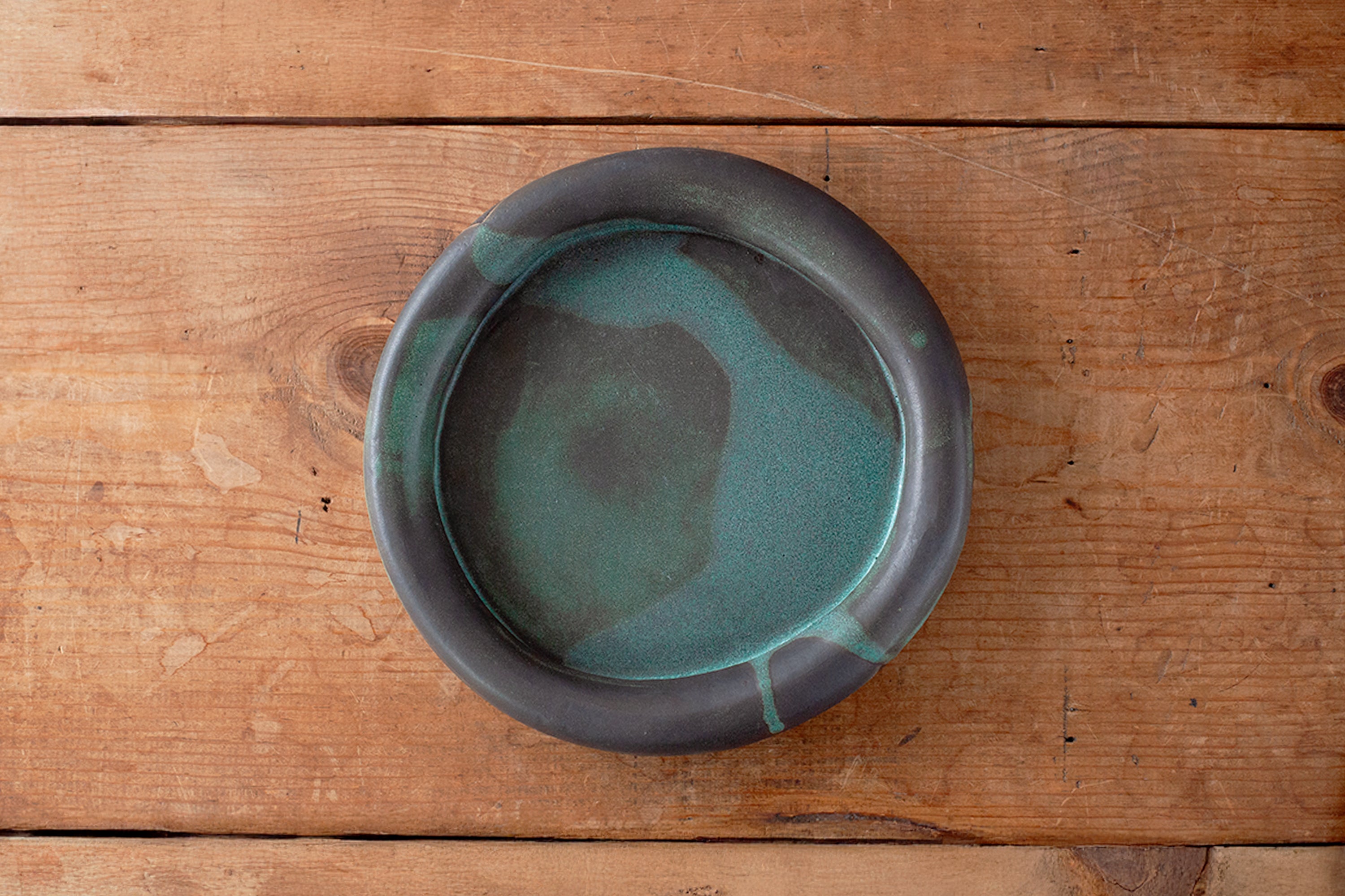 A round ceramic plate with a dark gray exterior and a teal-green glazed interior on a rustic wooden surface.