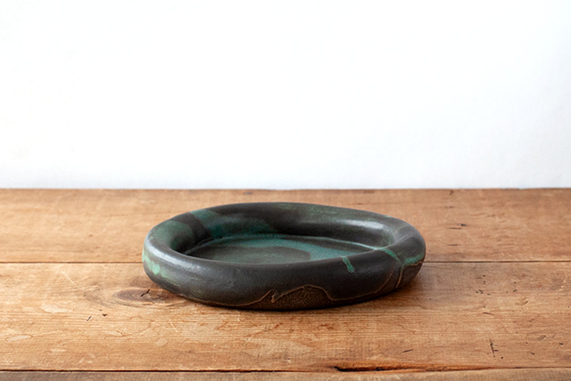 A round, rimmed ceramic plate with a dark green and black marbled glaze, resting on a rustic wooden surface against a plain white wall.