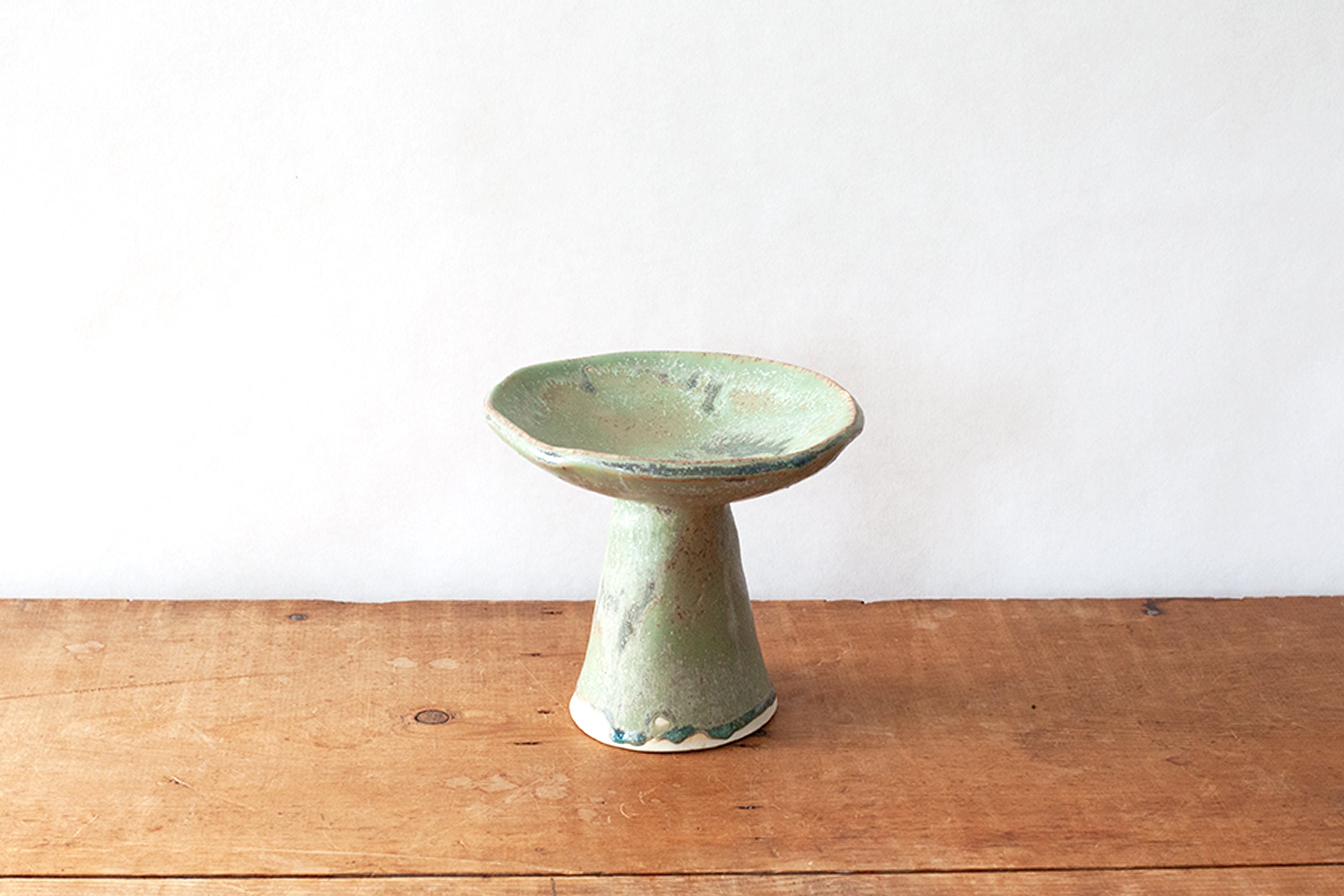 A handmade ceramic pedestal dish with a light green glaze and distressed brown edges, set on a rustic wooden table against a plain white wall.