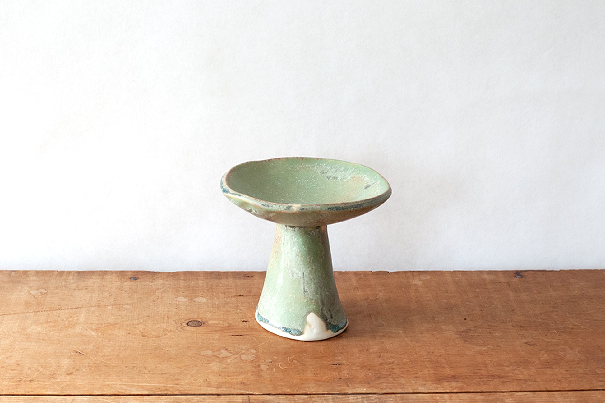 A small ceramic pedestal dish with a light green glaze and distressed finish, set on a rustic wooden surface against a plain white wall.