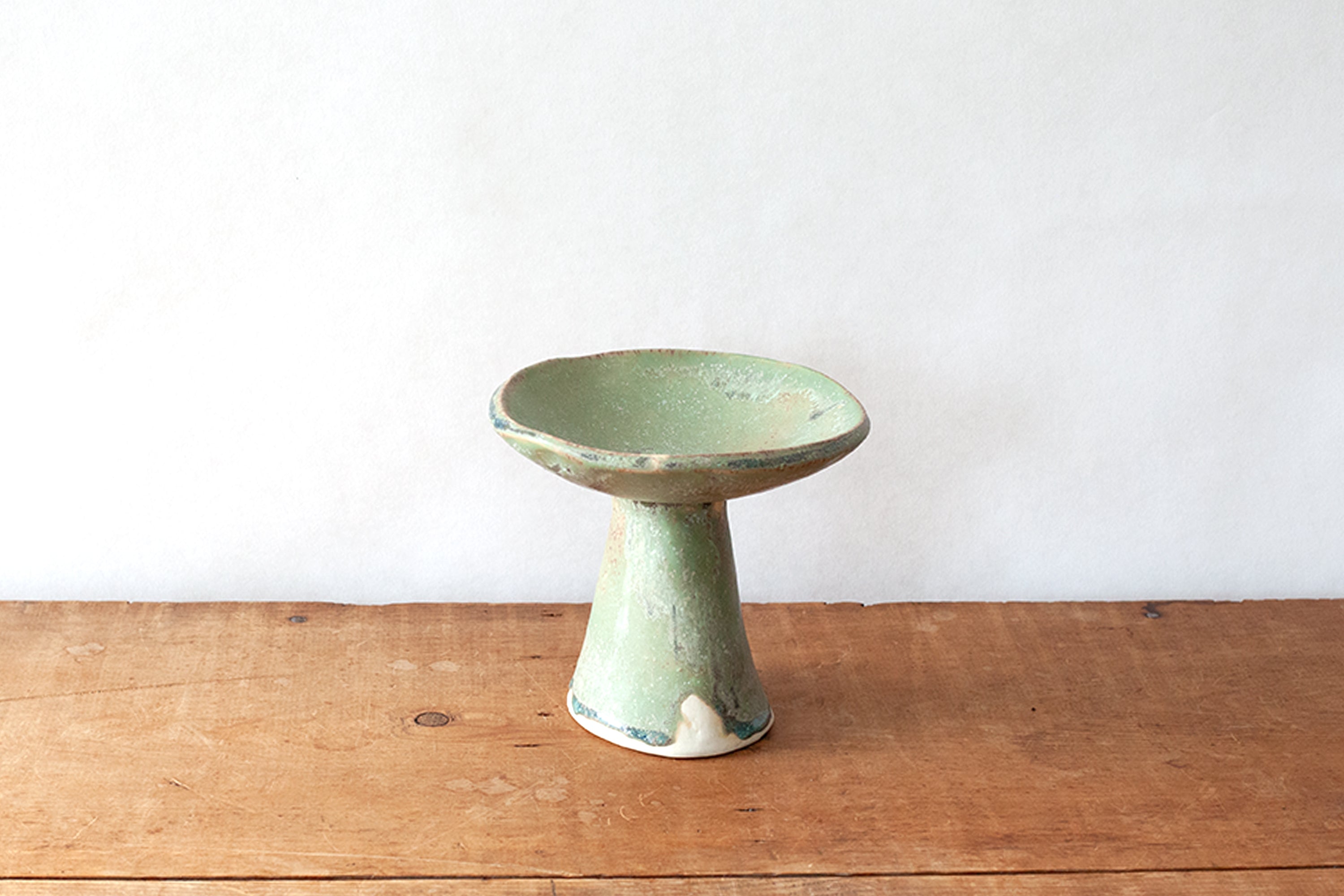 A small ceramic pedestal dish with a light green glaze and distressed finish, set on a rustic wooden surface against a plain white wall.