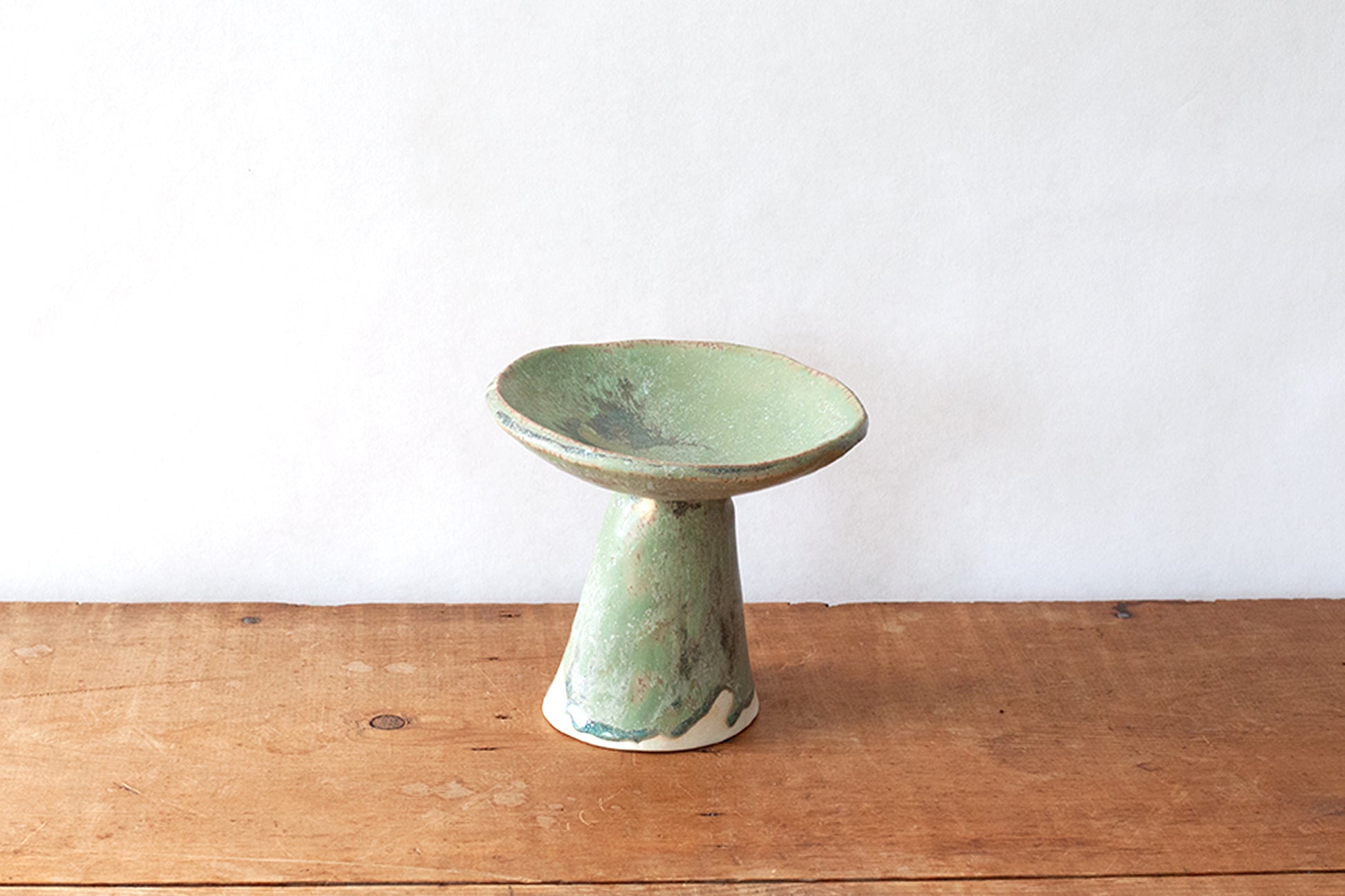 A handmade ceramic pedestal bowl with a light green glaze and earthy brown rim, set on a rustic wooden table against a plain white wall.