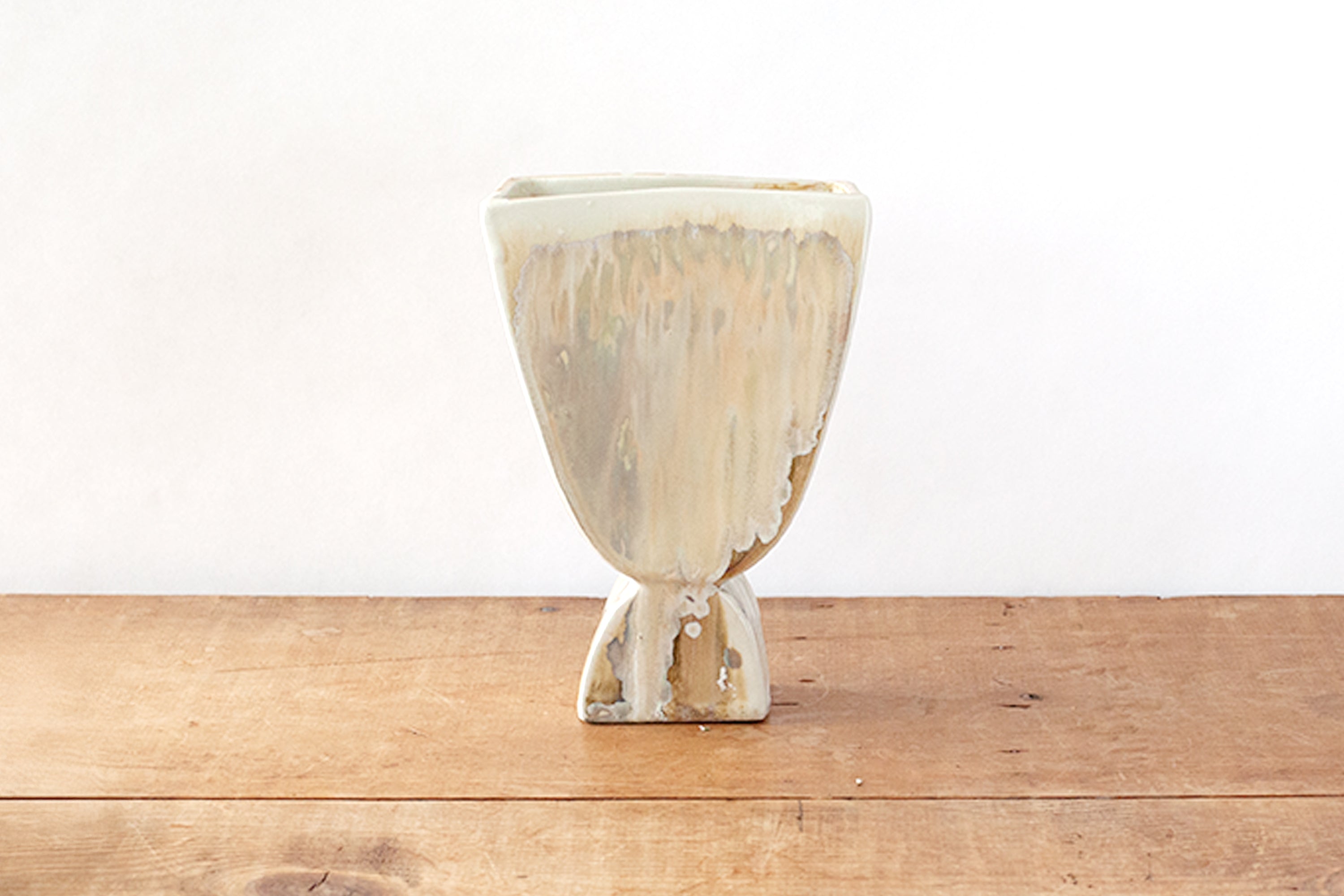 A ceramic cup or small vase with a textured, dripping glaze in cream and beige tones, standing on a rustic wooden surface against a plain white background.