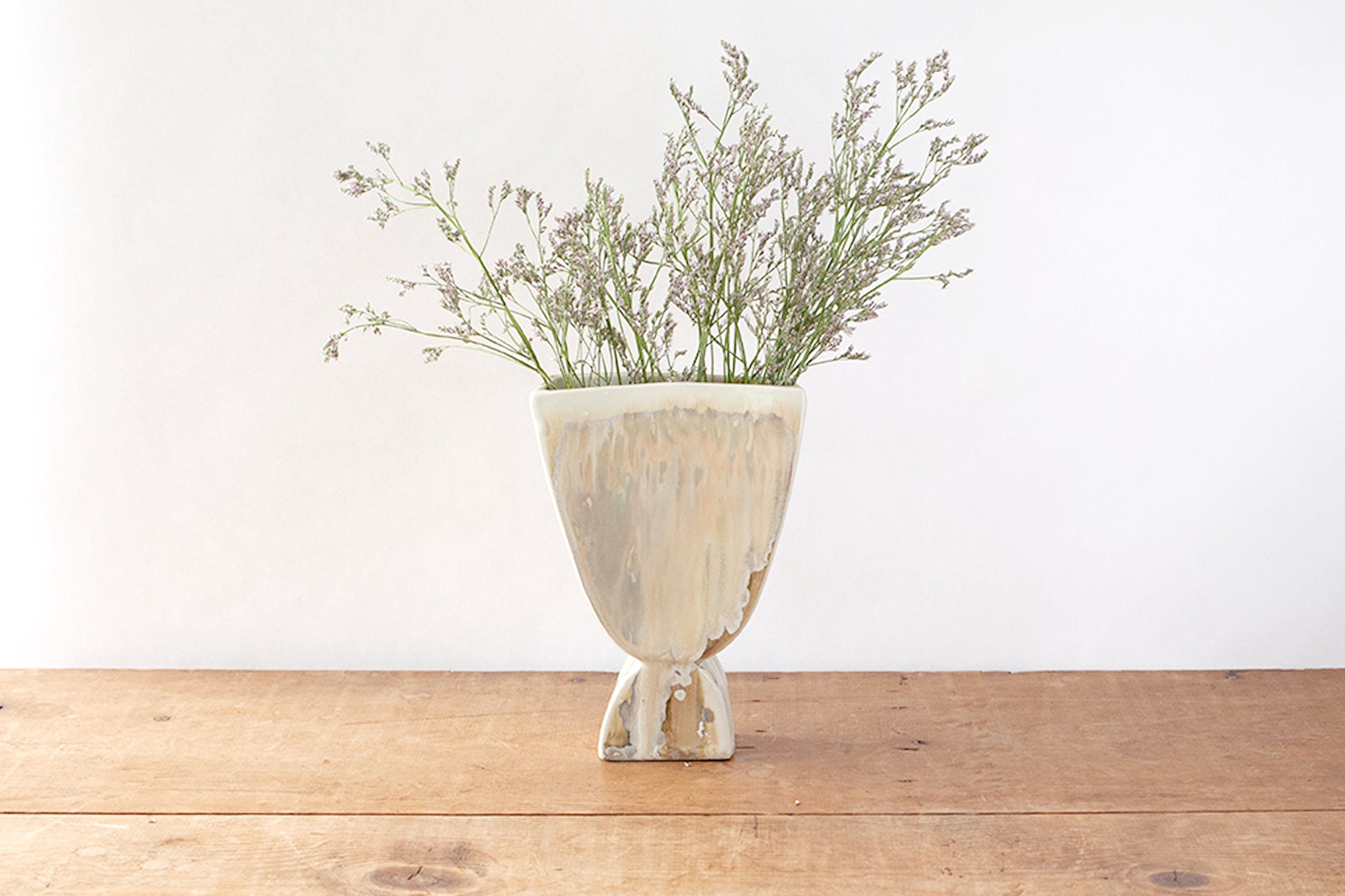 A rustic ceramic vase with a distressed cream and light brown glaze, holding dried greenish-brown grasses, set on a wooden tabletop against a plain white wall.
