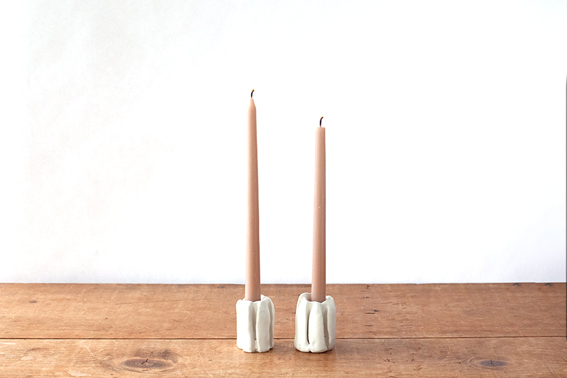 Two slim light pink taper candles standing in white ceramic candleholders on a rustic wooden tabletop against a plain white wall.