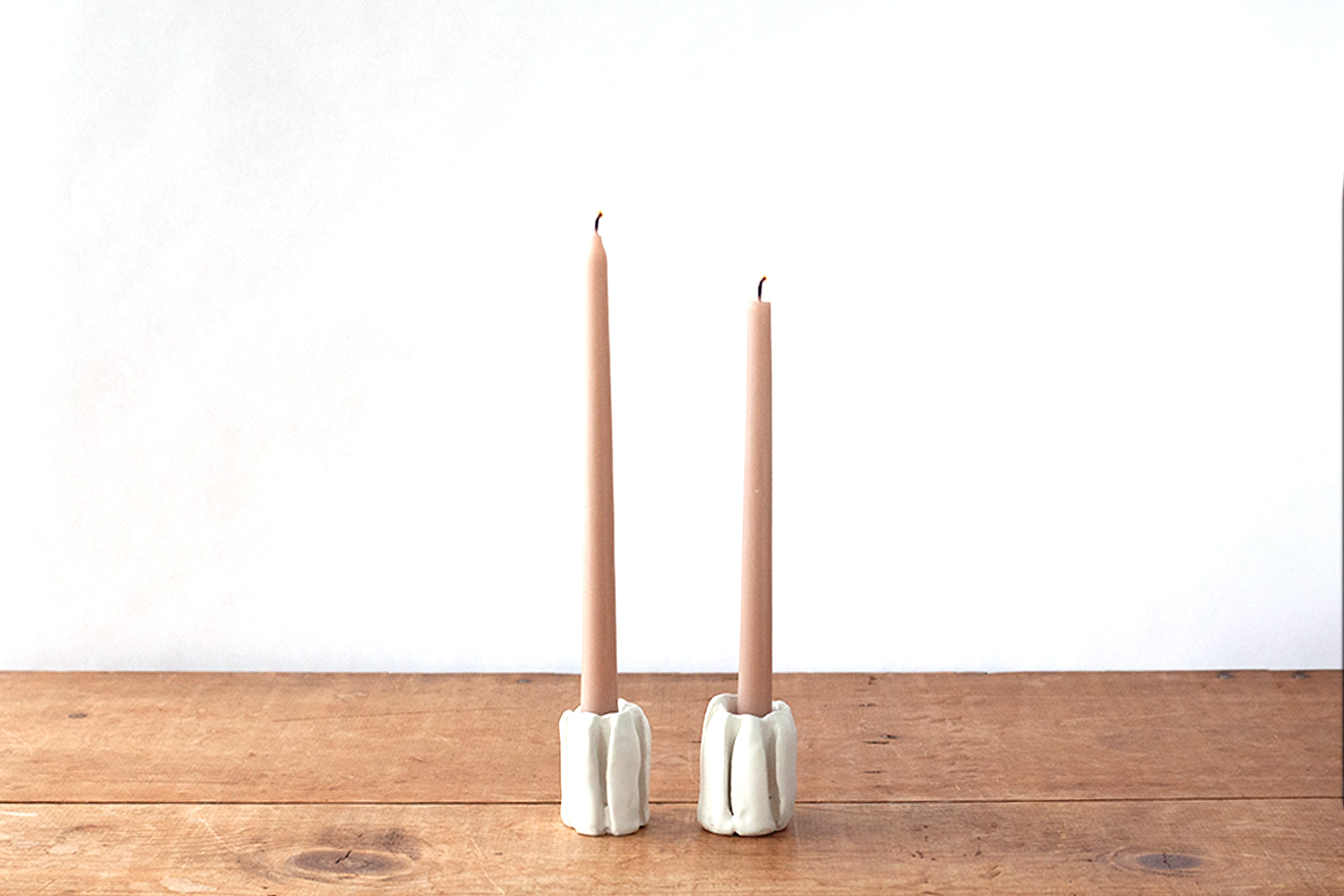 Two slim light pink taper candles standing in white ceramic candleholders on a rustic wooden tabletop against a plain white wall.