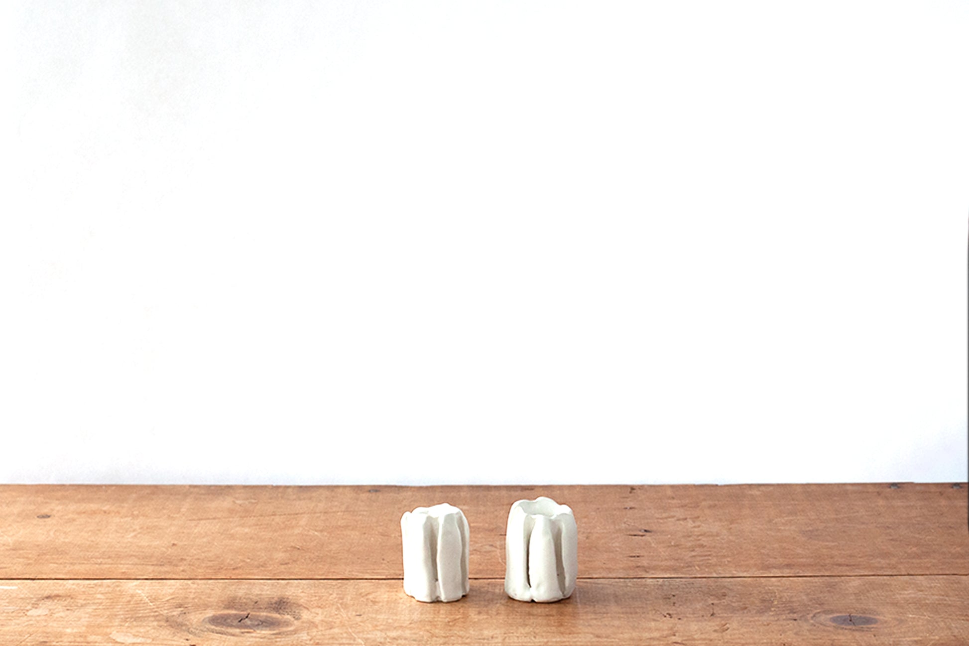 Two small white organically carved ribbed ceramic candleholders standing side by side on a rustic wooden tabletop against a plain white wall.