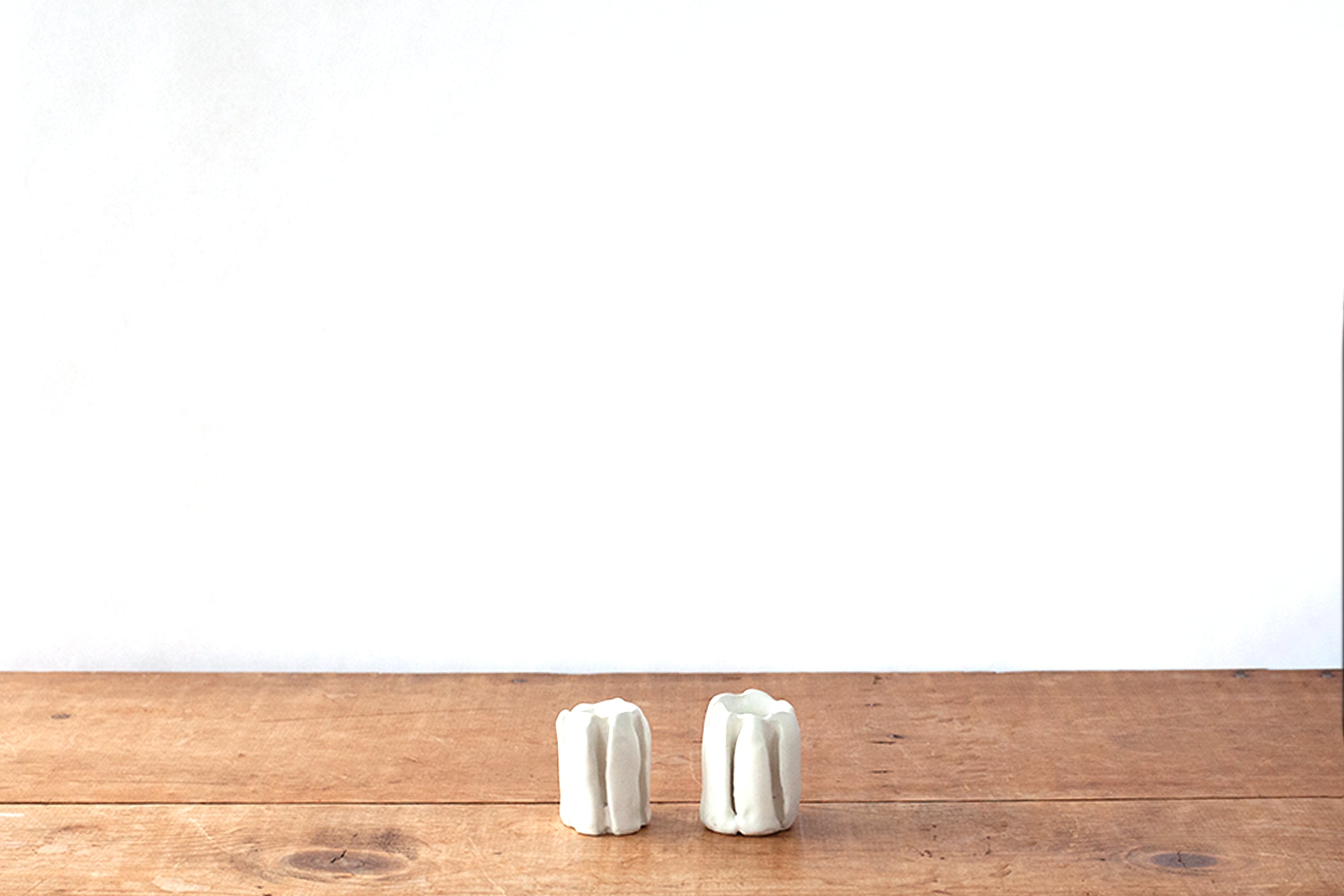 Two small white organically carved ribbed ceramic candleholders standing side by side on a rustic wooden tabletop against a plain white wall.