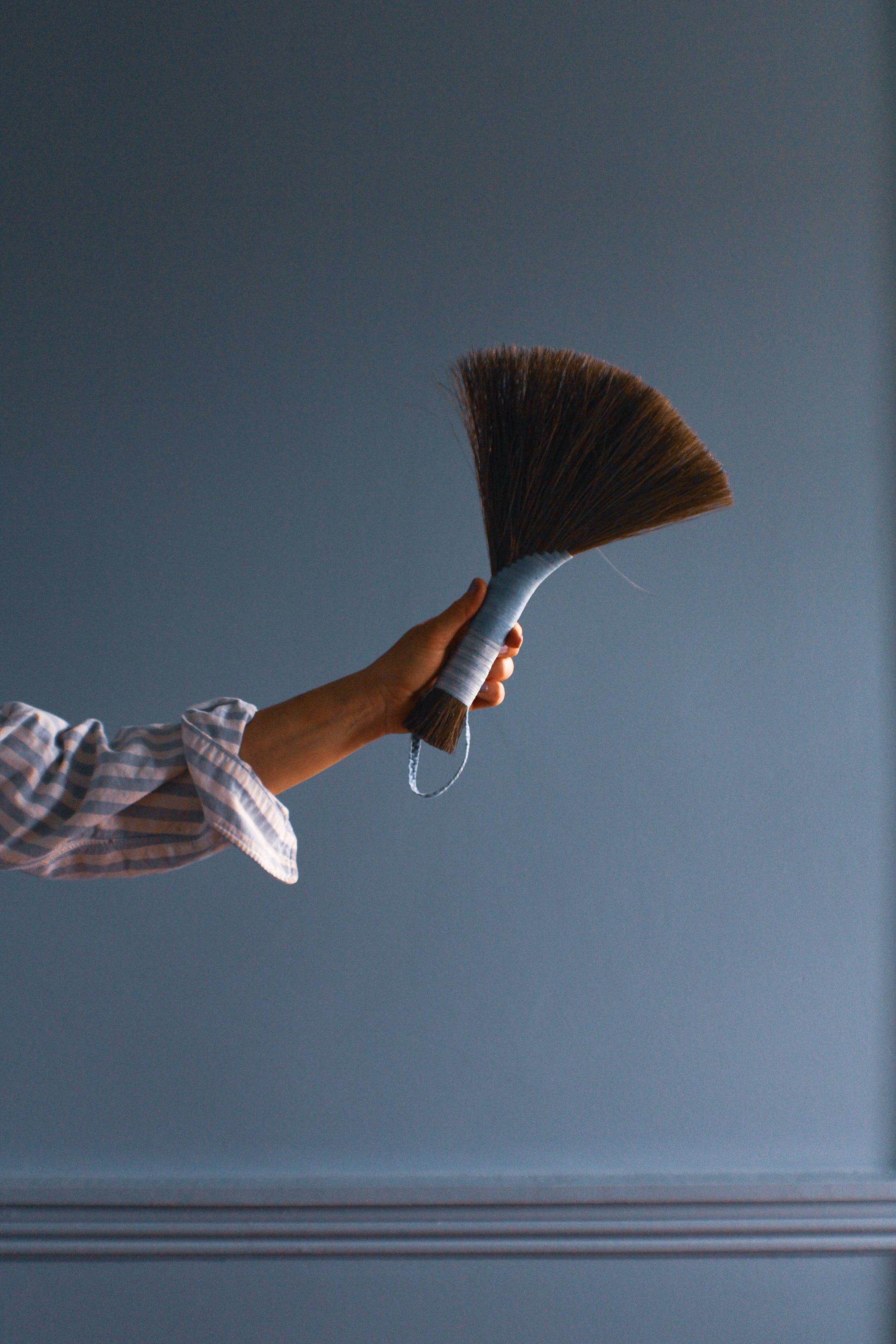 An arm extending out holding a hand broom against a plain blue wall