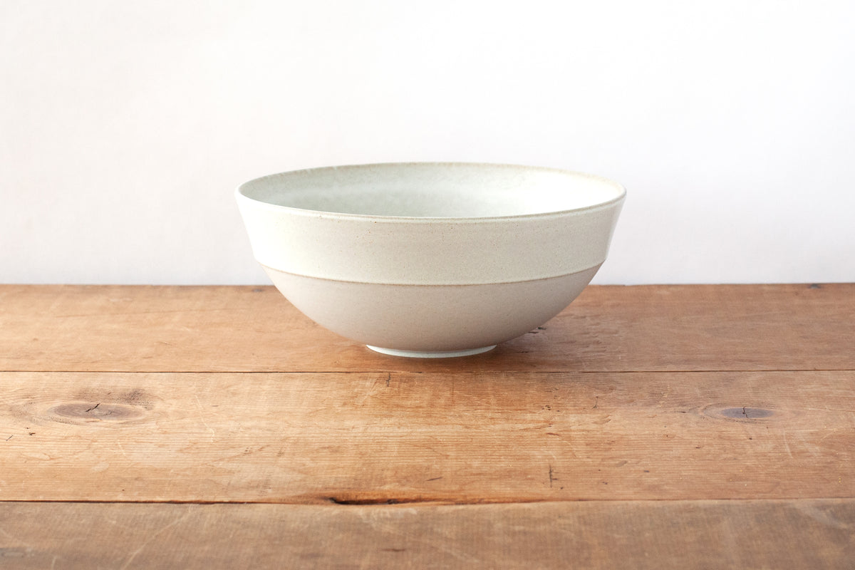 A light off-white ceramic bowl with a subtle horizontal ridged band, sitting on a rustic wooden tabletop against a plain white wall.