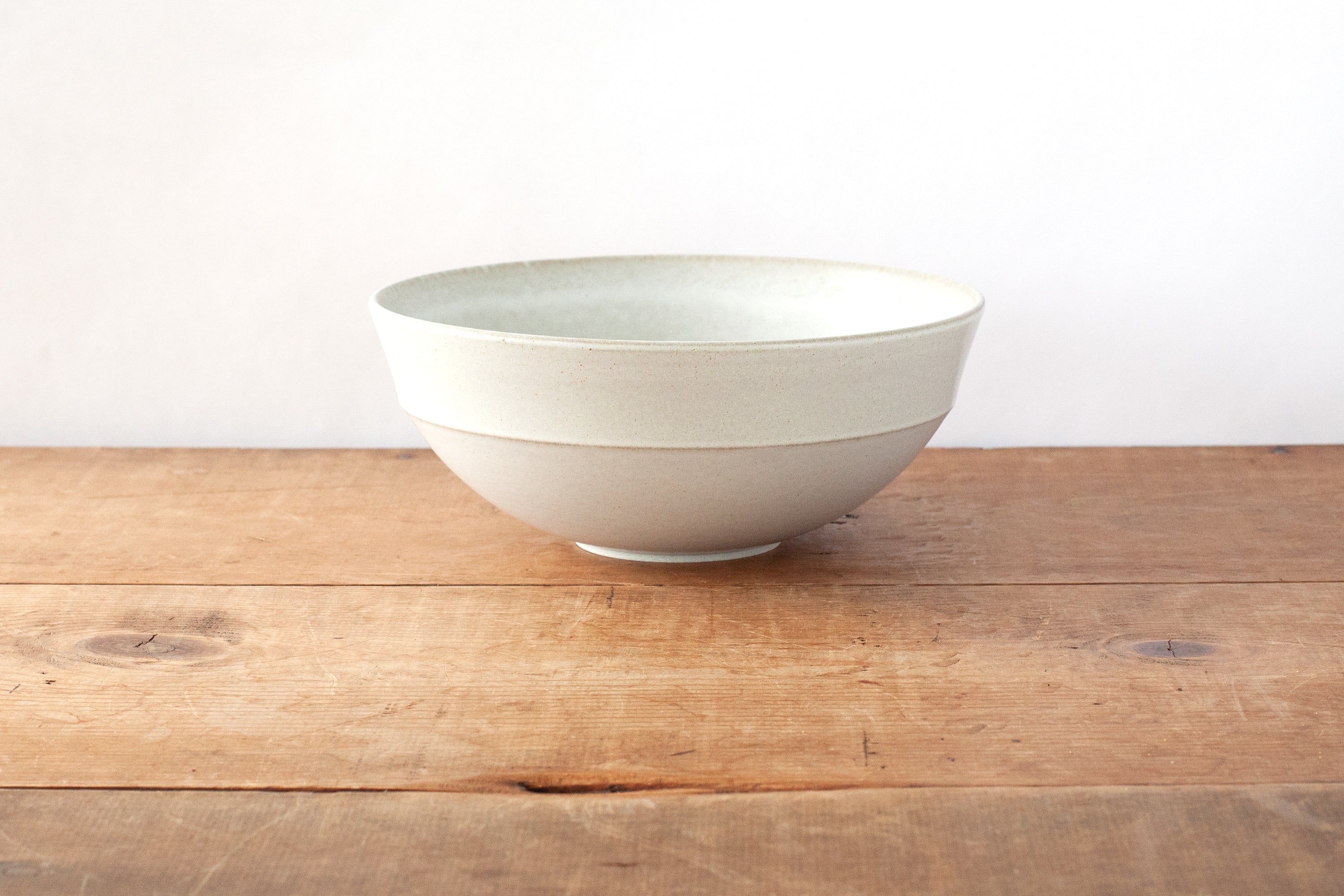 A light off-white ceramic bowl with a subtle horizontal ridged band, sitting on a rustic wooden tabletop against a plain white wall.